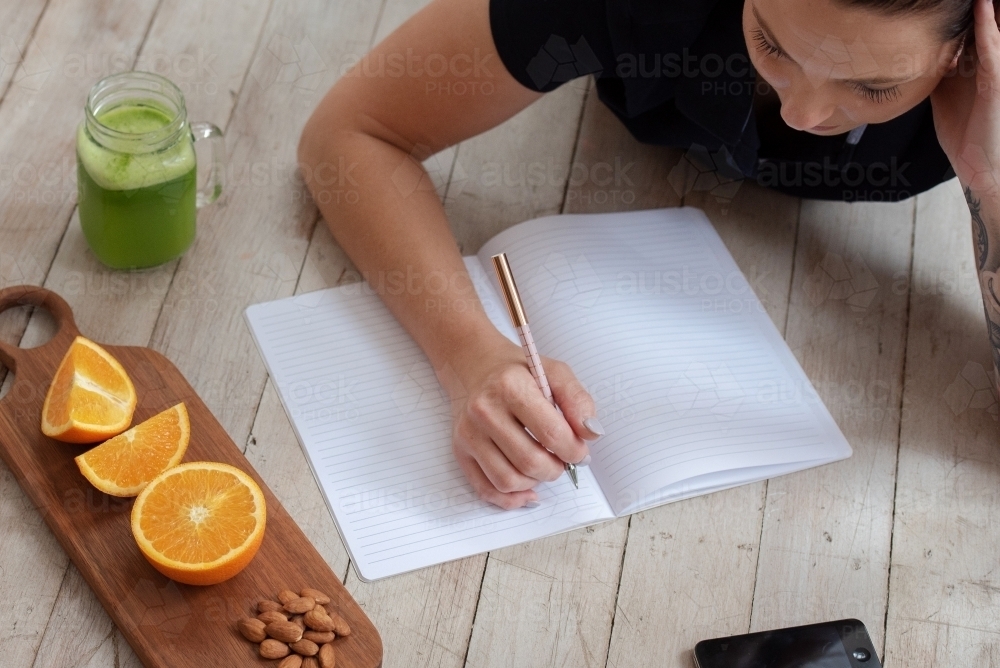 woman lying on floor journalling with green juice oranges almond healthy snack and phone - Australian Stock Image