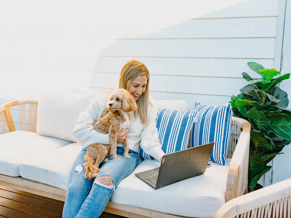 Woman on her laptop while holding a dog on the couch. - Australian Stock Image