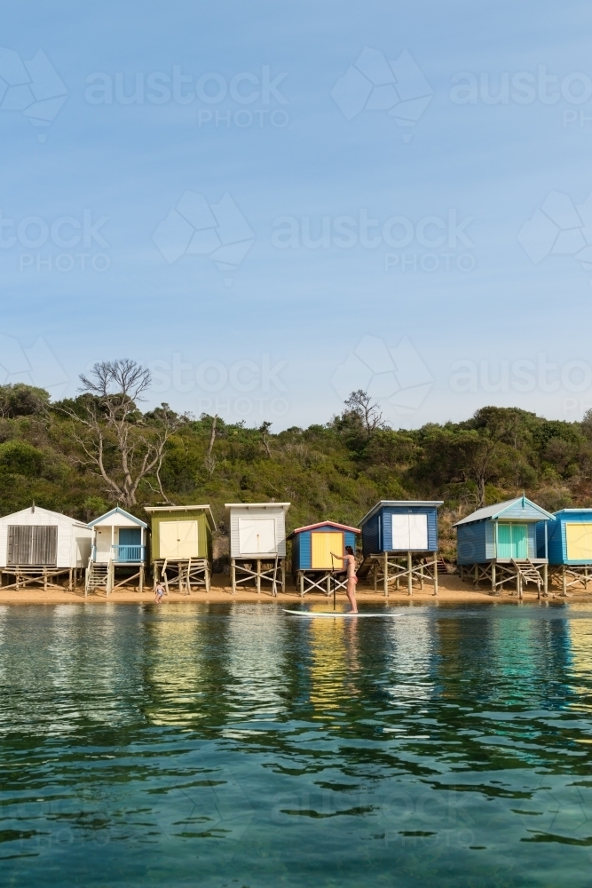 Woman on stand up paddle board in front of  beach huts - Australian Stock Image