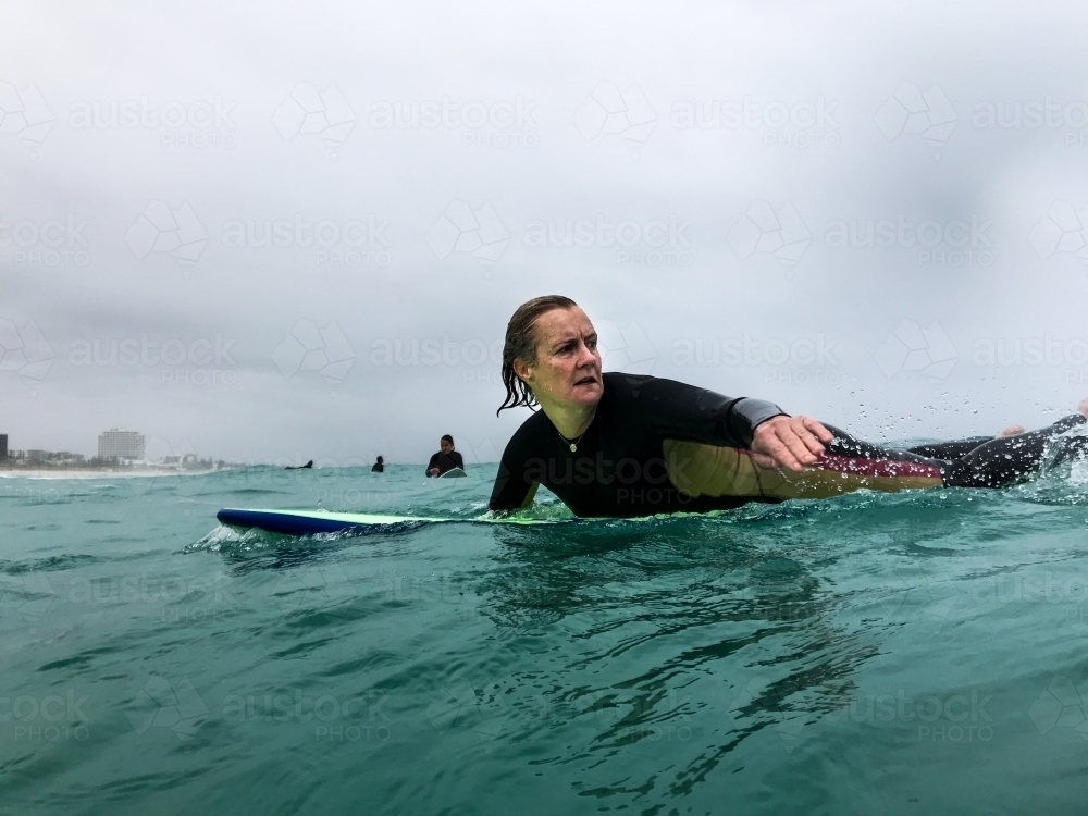 woman paddling on surfboard looking back into surf on overcast day - Australian Stock Image
