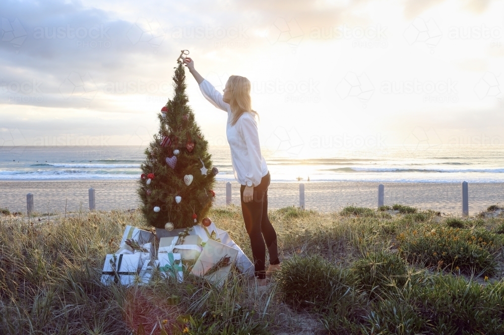 Woman placing decoration on top of Christmas tree on beach at sunrise - Australian Stock Image