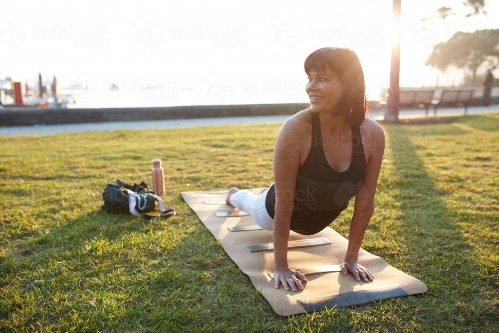Woman practising yoga at sunrise in the park by harbour - Australian Stock Image