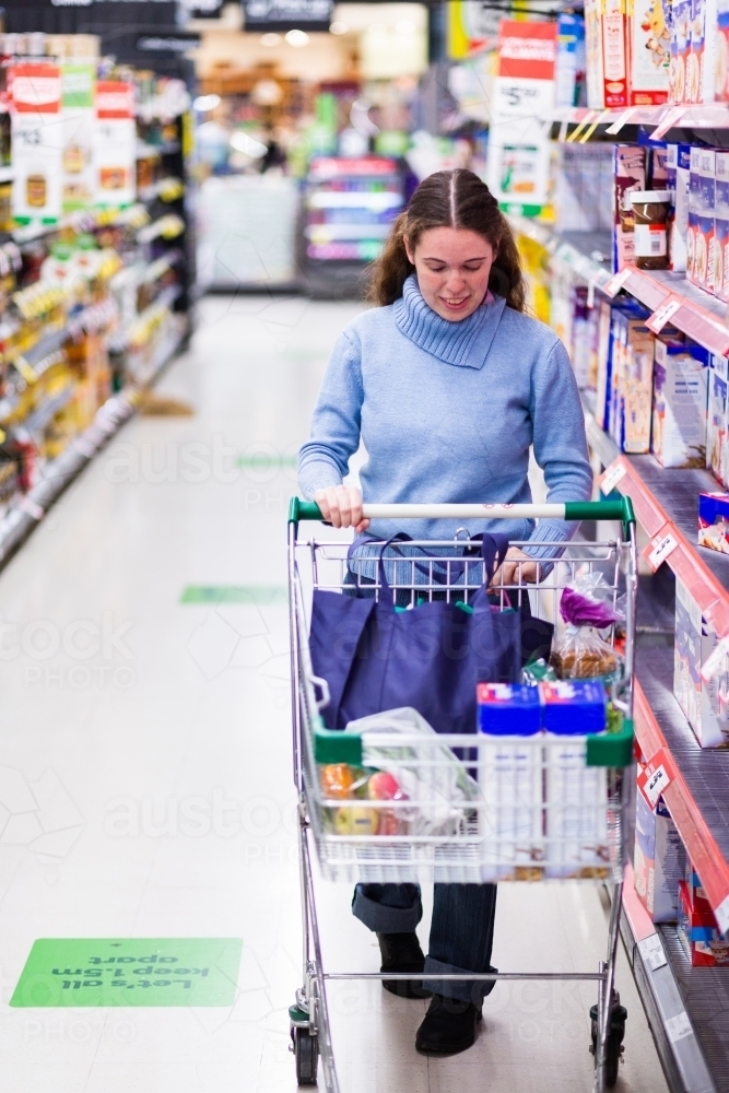 Woman pushing shopping trolley through aisles of supermarket doing household shop - Australian Stock Image