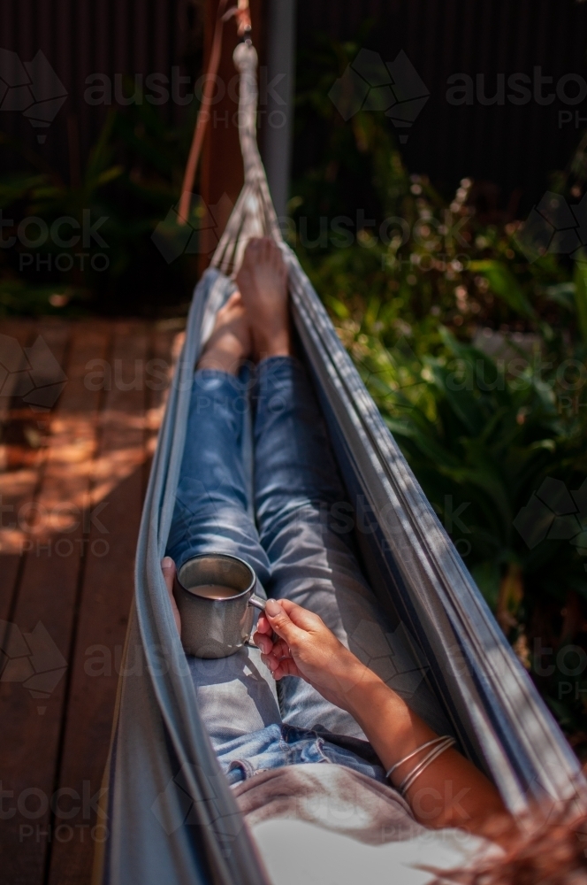 Woman relaxing in hammock holding hot drink in mug - Australian Stock Image