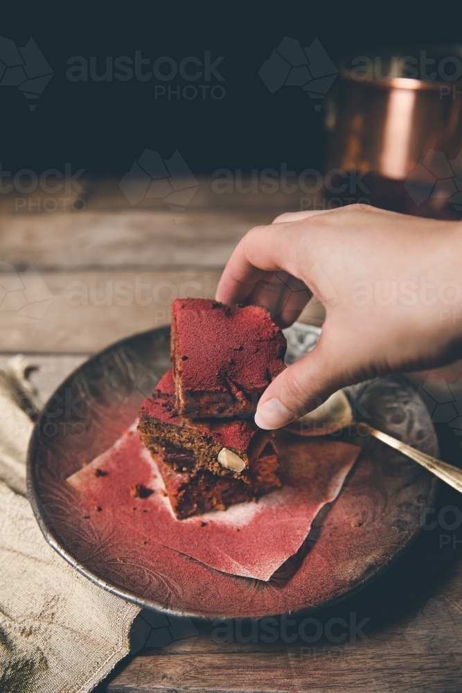 Woman's hand picks up a red dusted chocolate brownie on timber surface with dark background - Australian Stock Image