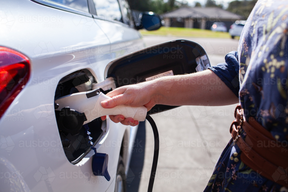 Woman’s hand plugging in hybrid car to charge EV battery in suburban driveway - Australian Stock Image