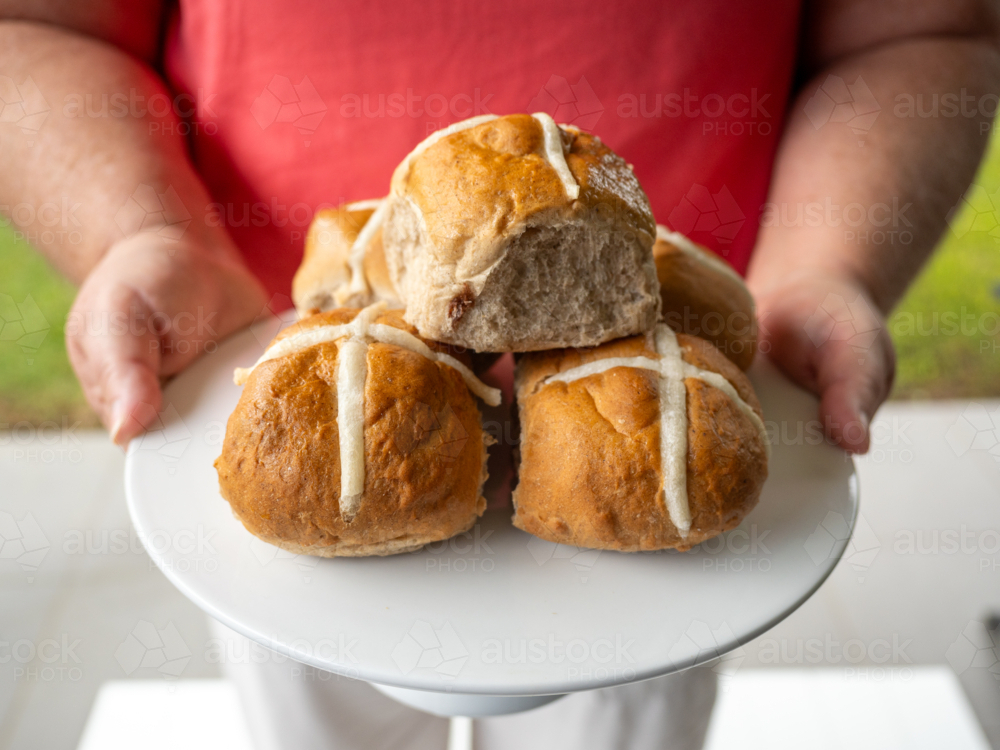 Woman's hands holding white plate piled with hot cross buns in backyard - Australian Stock Image
