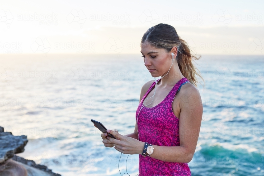 Woman selecting music track whilst exercising on coastline - Australian Stock Image