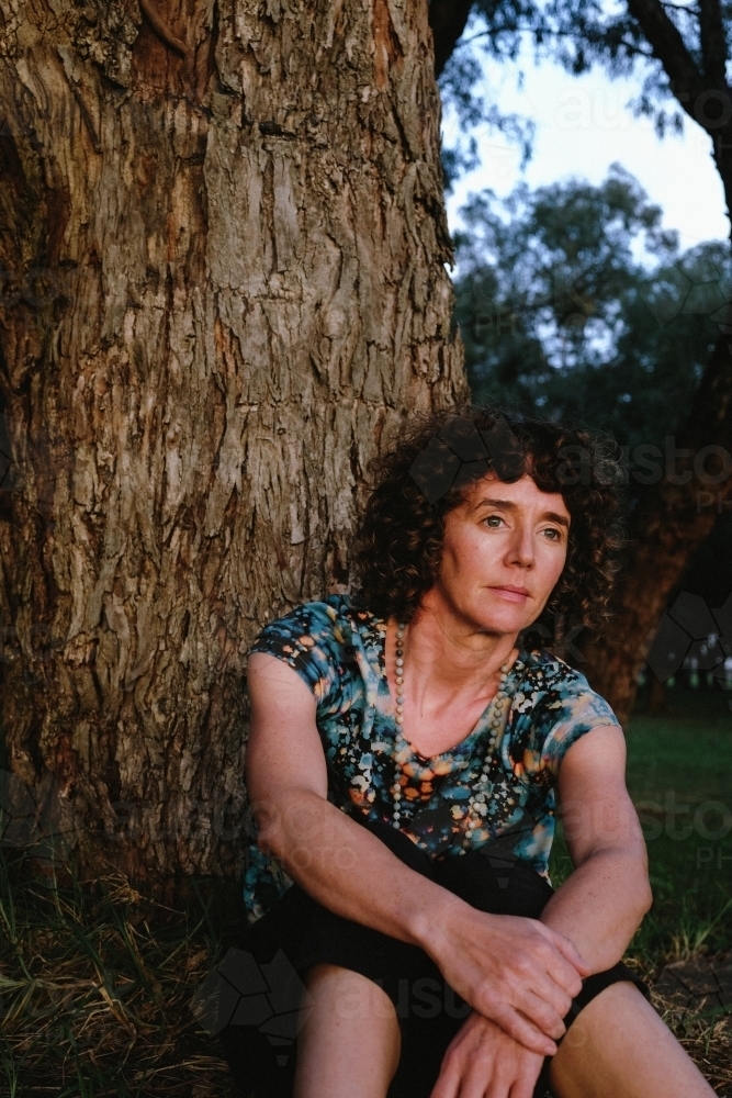 Woman sitting against trunk of tree in golden light - Australian Stock Image