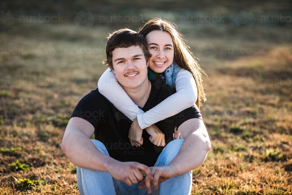 Woman sitting behind boyfriend with arms wrapped around him smiling - Australian Stock Image