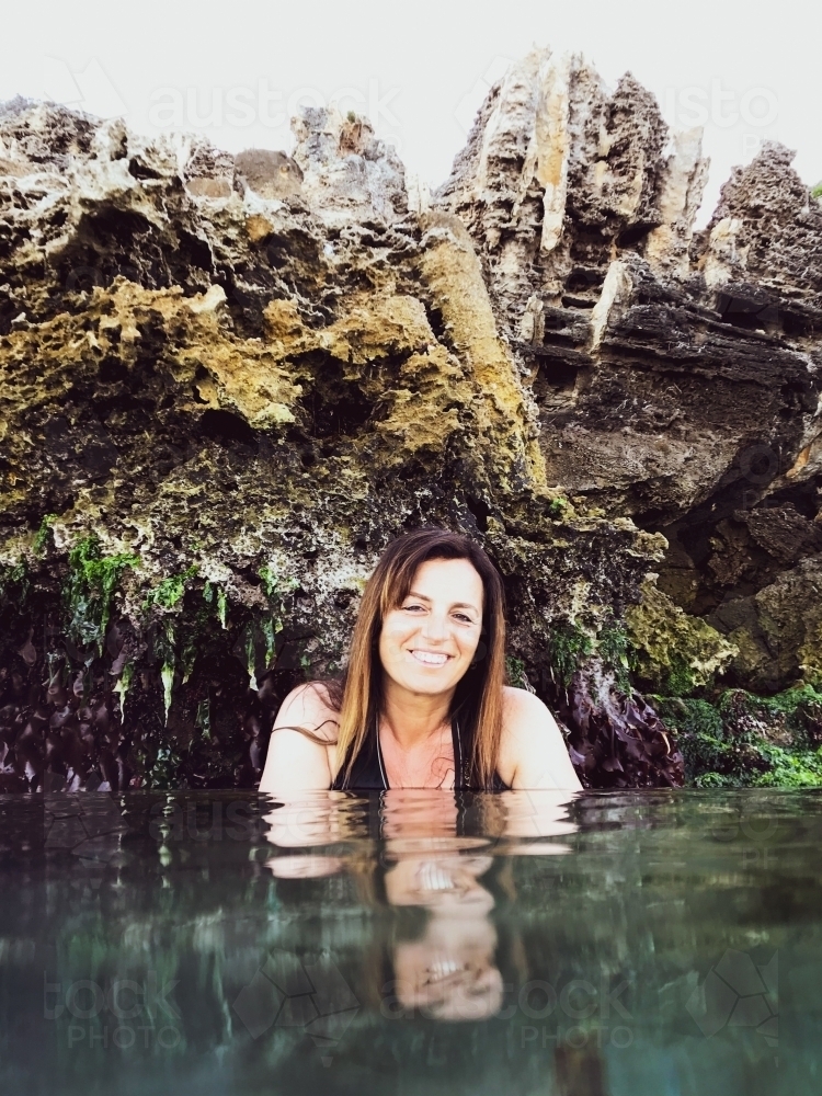 Woman sitting in glassy ocean and cupping water with coastal limestone rocks in background - Australian Stock Image