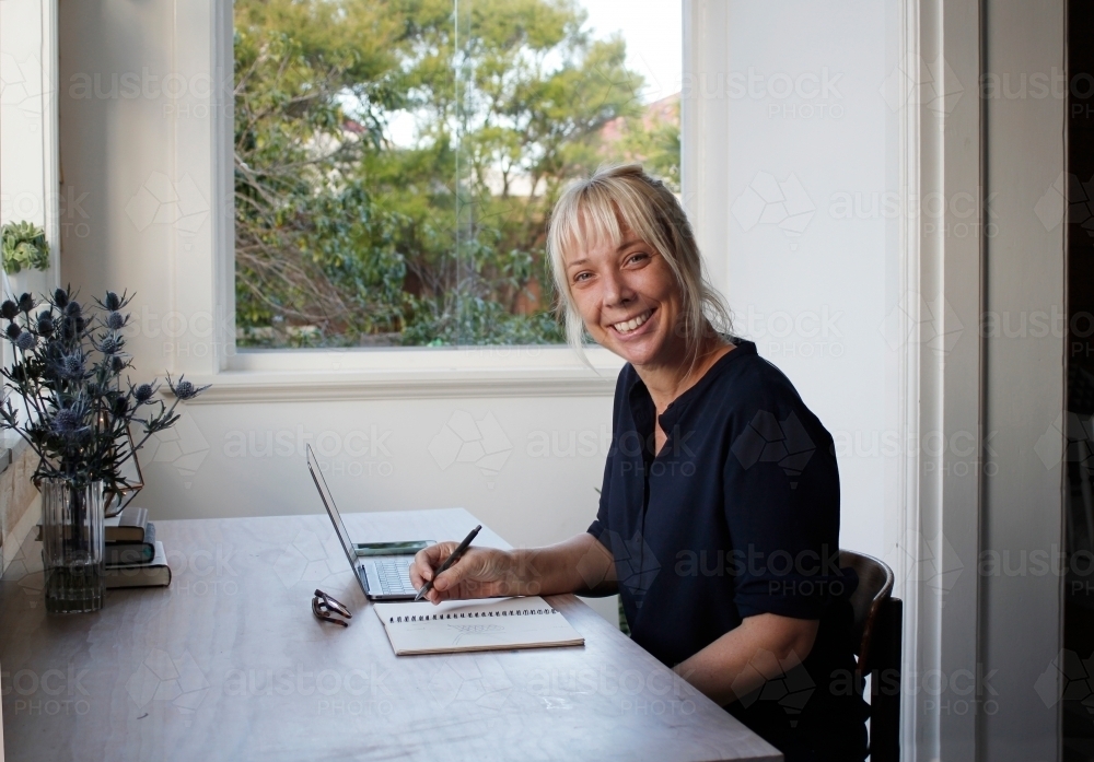 Woman sitting in sun room office working with laptop and notepad - Australian Stock Image