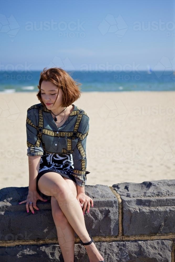 Woman sitting on a bluestone wall with the beach, sand and sea behind her - Australian Stock Image