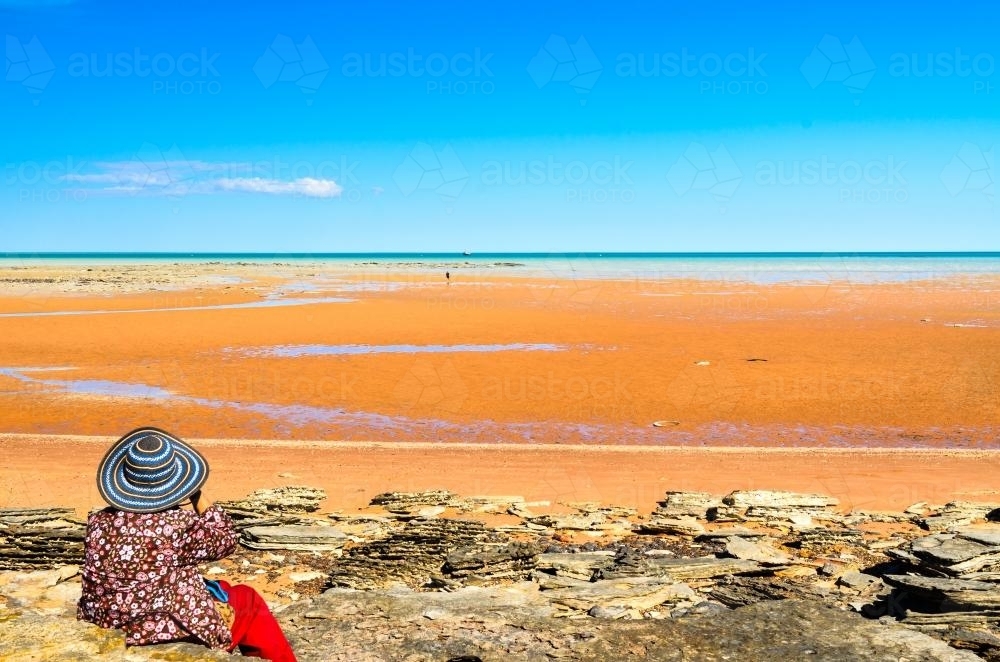 Woman sitting on beach at low tide looking out at sea - Australian Stock Image