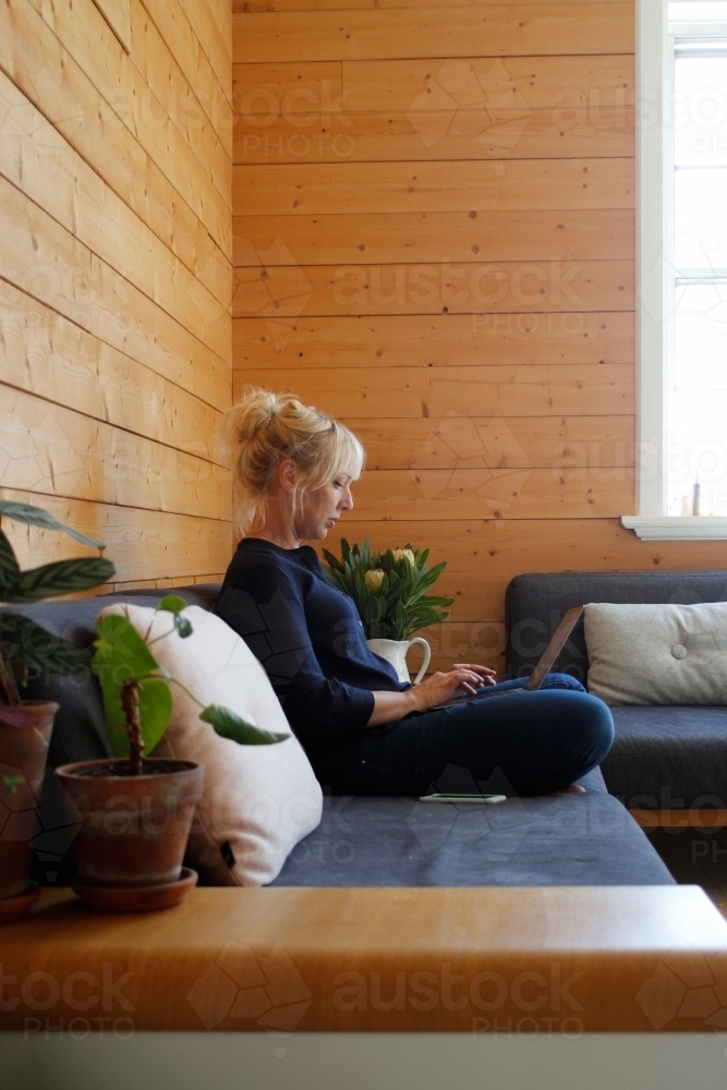 Woman sitting on couch working on laptop with timber wall background - Australian Stock Image
