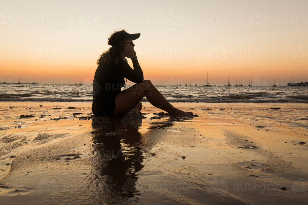 Woman sitting on shoreline listening to music on headphones at sunset - Australian Stock Image