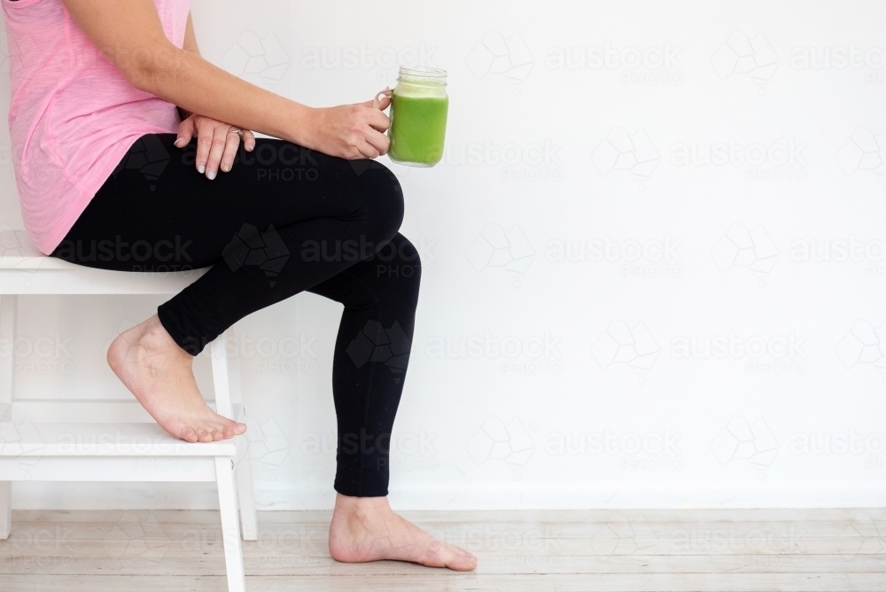 woman sitting on stool in workout gear holding green juice - Australian Stock Image