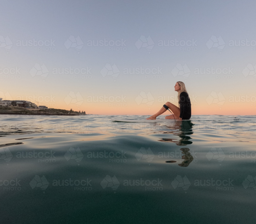 Woman sitting on surfboard floating on glassy water with leg up on board and sunrise sky - Australian Stock Image