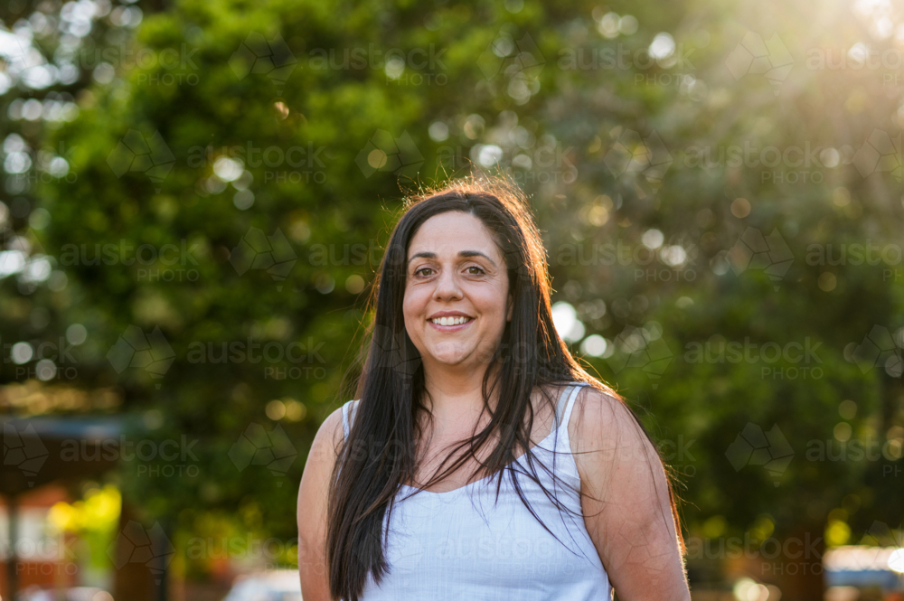 Woman smiles while standing in a park as sunlight shines through trees around her. - Australian Stock Image