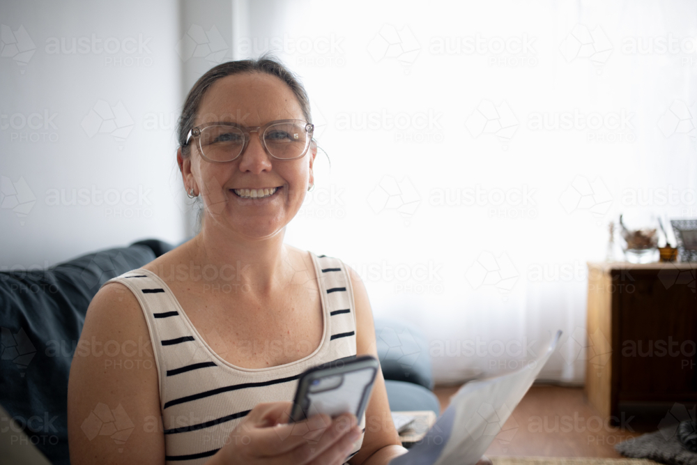 Woman smiling at camera, using smartphone and reading document at home - Australian Stock Image