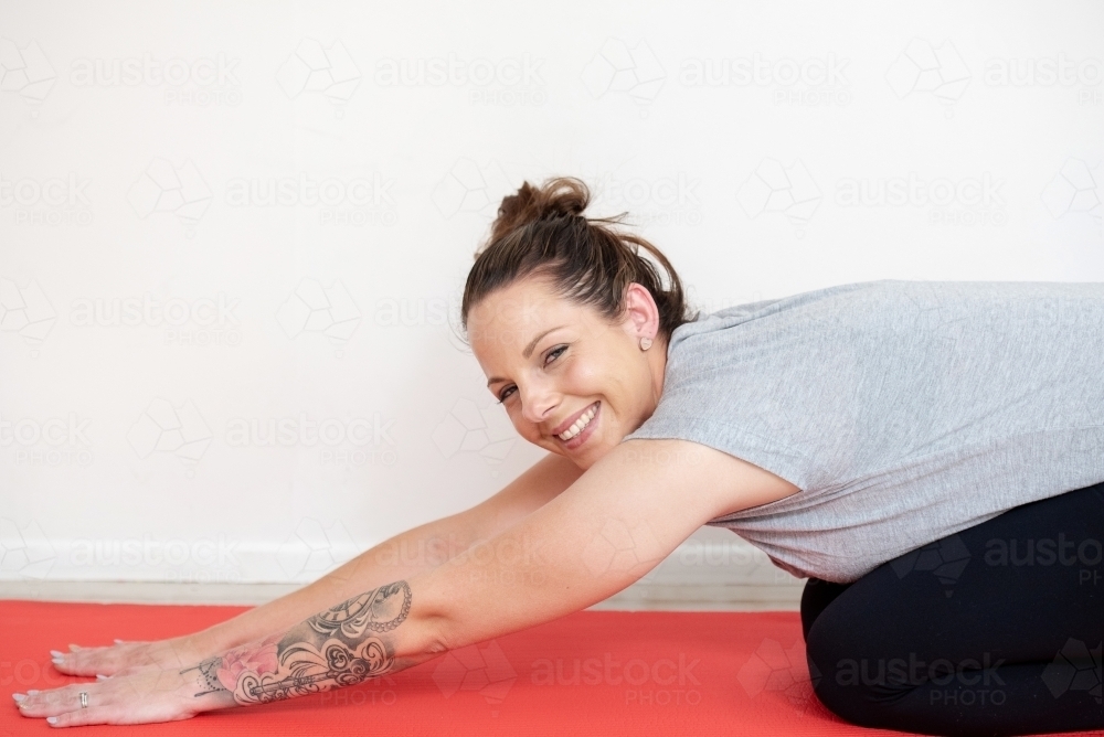 woman smiling at camera while doing child's pose on yoga mat in studio - Australian Stock Image
