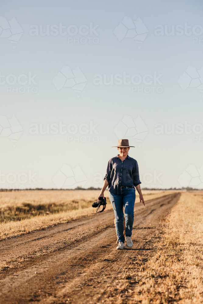 Woman smiling walking along a road carrying camera - Australian Stock Image
