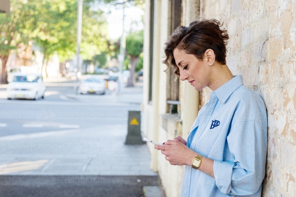 woman standing against brick wall using mobile phone - Australian Stock Image