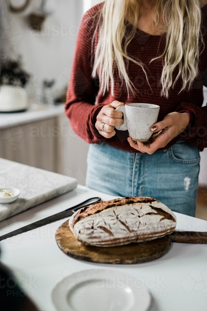 Woman standing behind kitchen bench holding a cup of coffee - Australian Stock Image