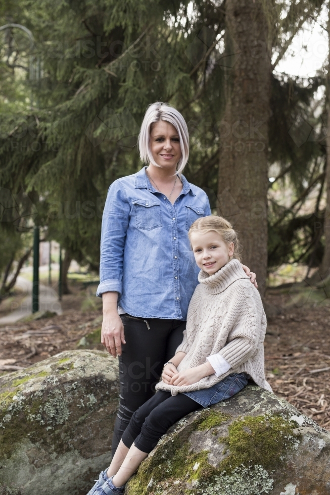 Woman standing beside young girl sitting on a rock outdoors - Australian Stock Image