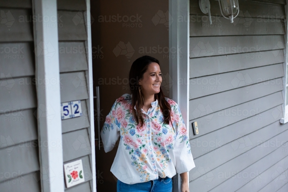 Woman standing by the open door of her house looking outside smiling - Australian Stock Image