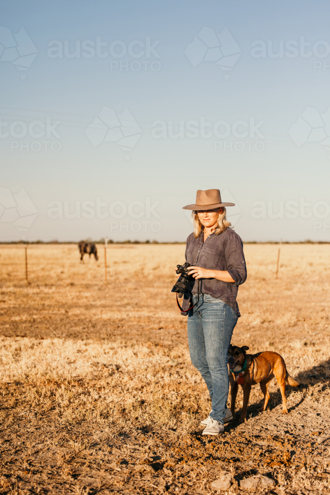 Woman standing in a dusty and dry grassy farm paddock holding a camera - Australian Stock Image