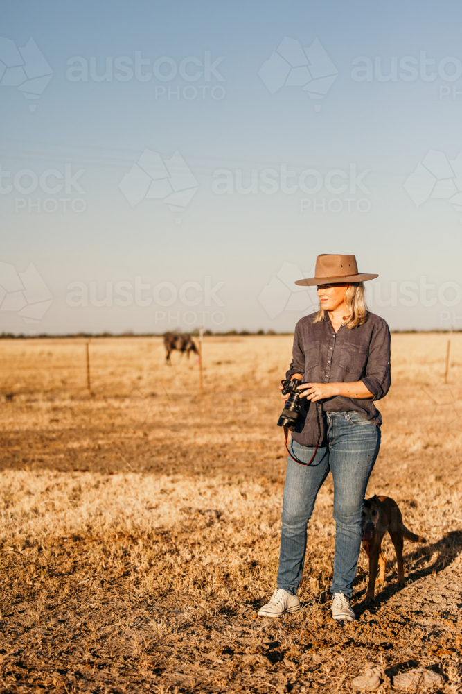 Woman standing in a dusty and dry grassy farm paddock holding a camera - Australian Stock Image