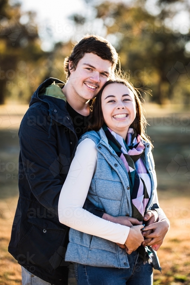Woman standing laughing in front of boyfriend arms wrapped around her holding hands - Australian Stock Image