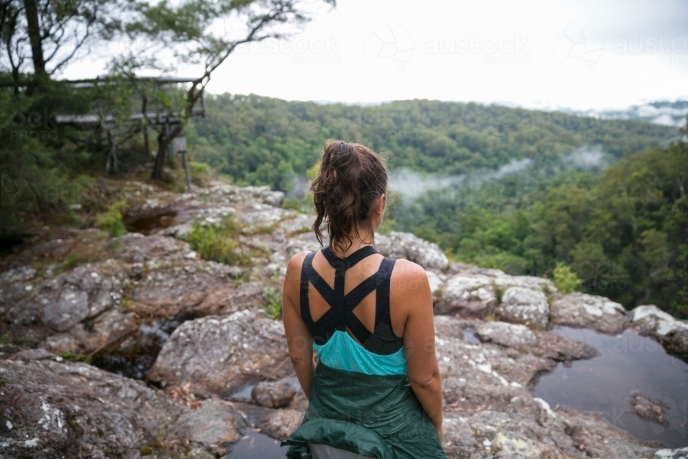 Woman standing on cliffs edge looking out at the rainforest view below - Australian Stock Image
