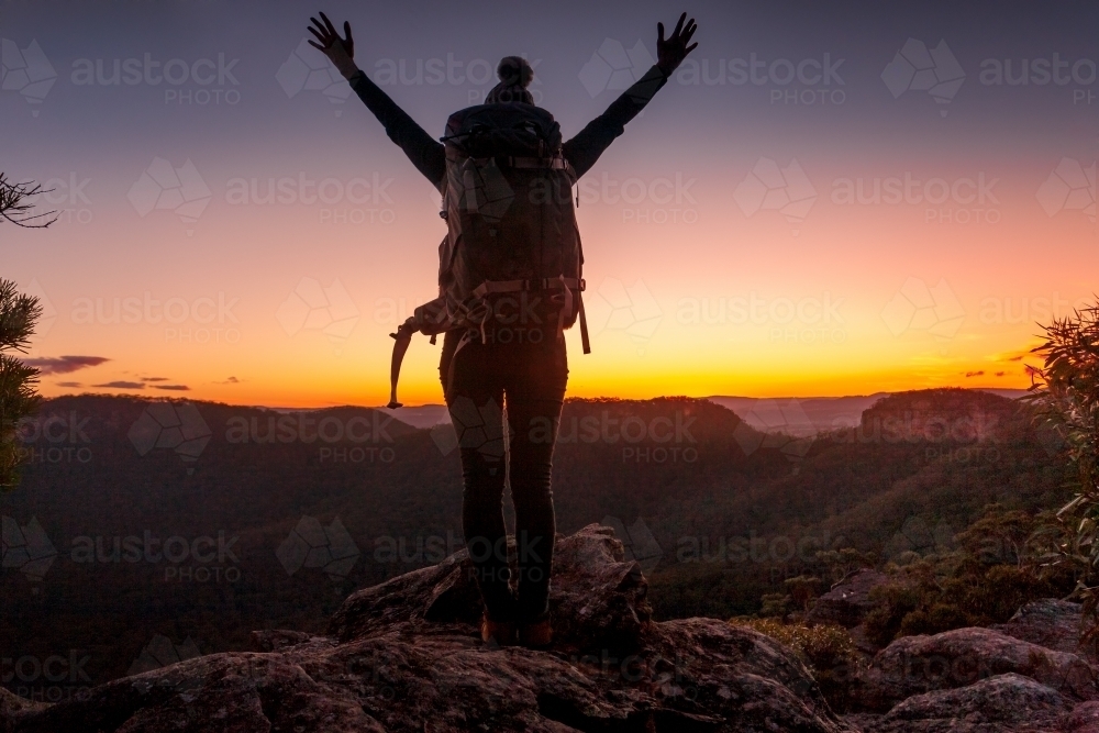 Woman standing on top of mountain peak views to escarpment and valleys, hands in air with feelings o - Australian Stock Image