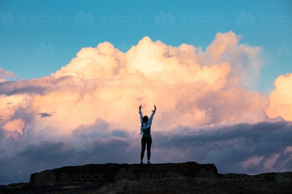 Woman standing with arms up on rock against a dramatic cloud backdrop - Australian Stock Image