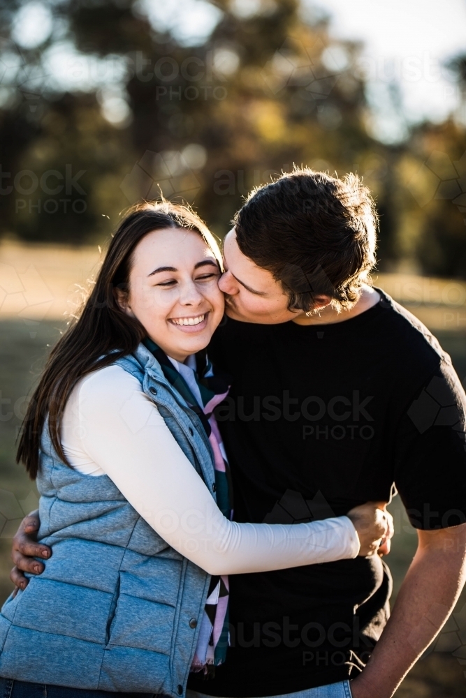 Woman standing with arms wrapped around boyfriend smiling while he kisses her on cheek - Australian Stock Image