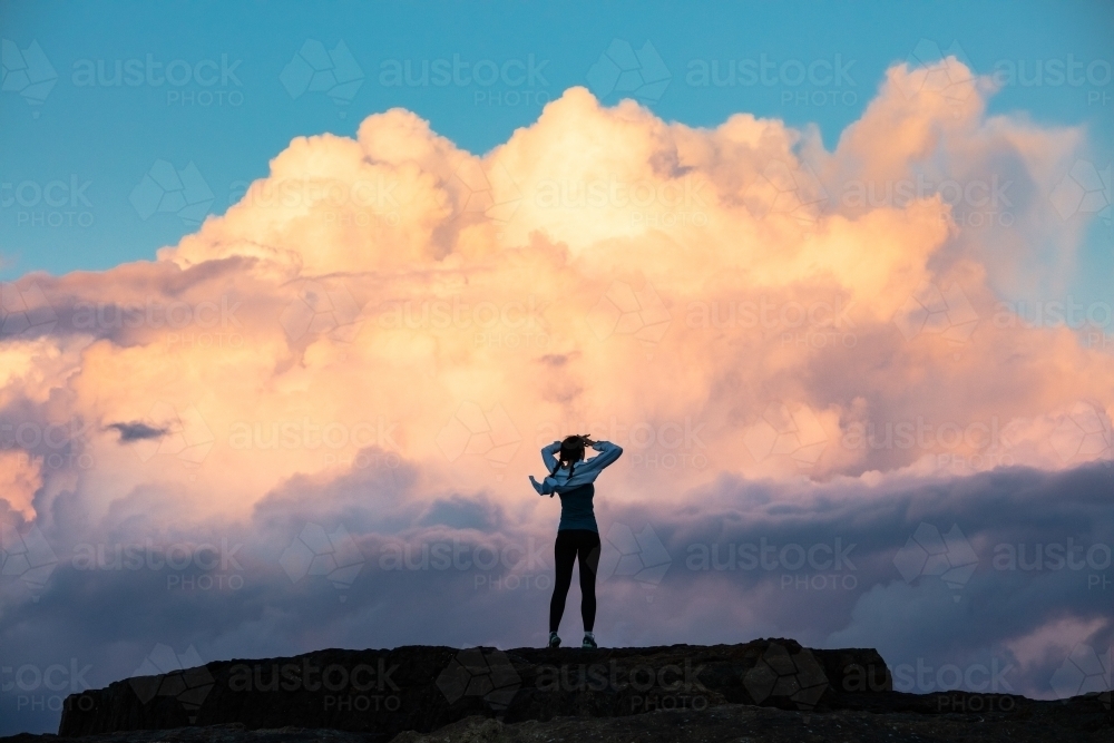 Woman standing with hands on head on rock against a dramatic cloud backdrop - Australian Stock Image