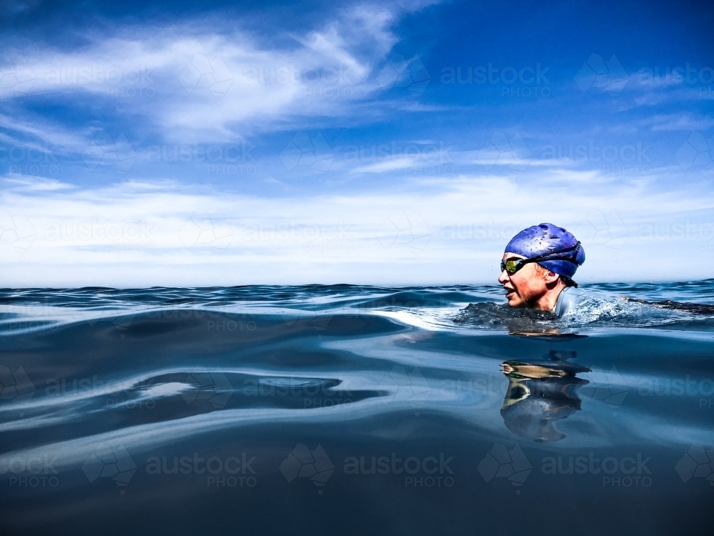 Woman swimming breastroke in cap and goggles in glassy ocean - Australian Stock Image