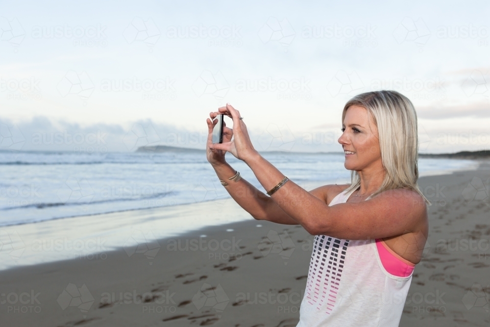 woman taking a photo at the beach on her phone - Australian Stock Image
