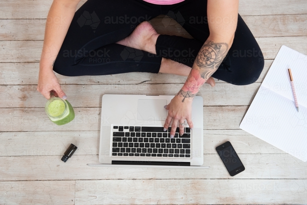 woman typing on computer with green juice essential oils journal pen phone in workout clothes - Australian Stock Image