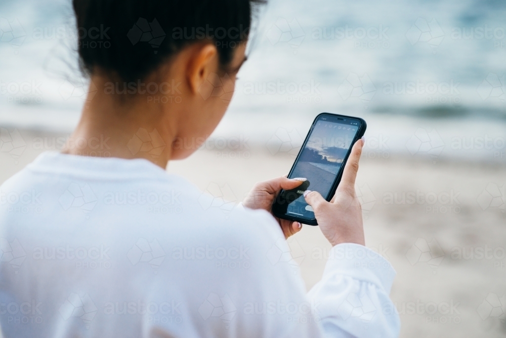 woman using phone at beach to take a sunset photo - Australian Stock Image