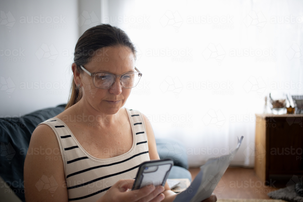 Woman using smartphone and reading document at home - Australian Stock Image