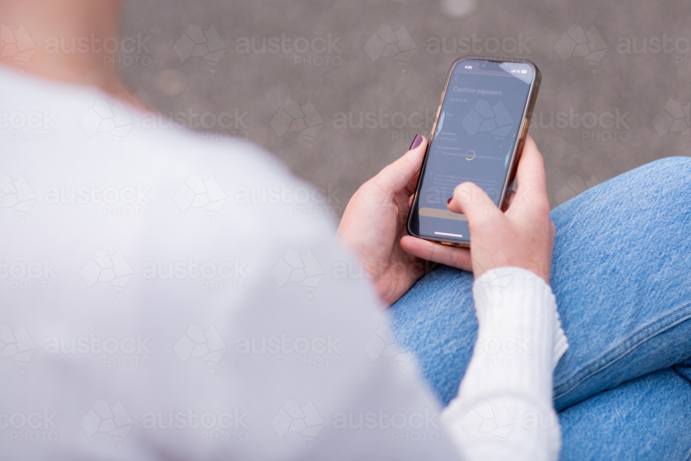 Woman using smartphone device outdoors while seated in jeans and white ribbed sweater - Australian Stock Image