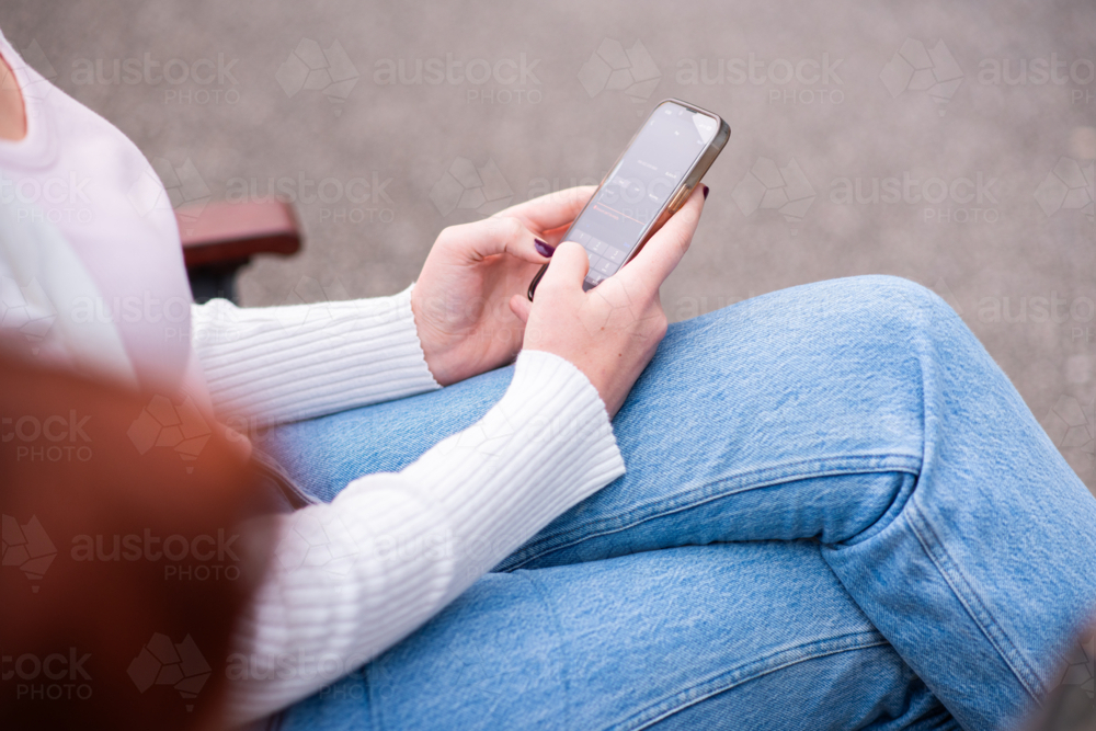 Woman using smartphone outdoors while seated in jeans and white ribbed sweater - Australian Stock Image