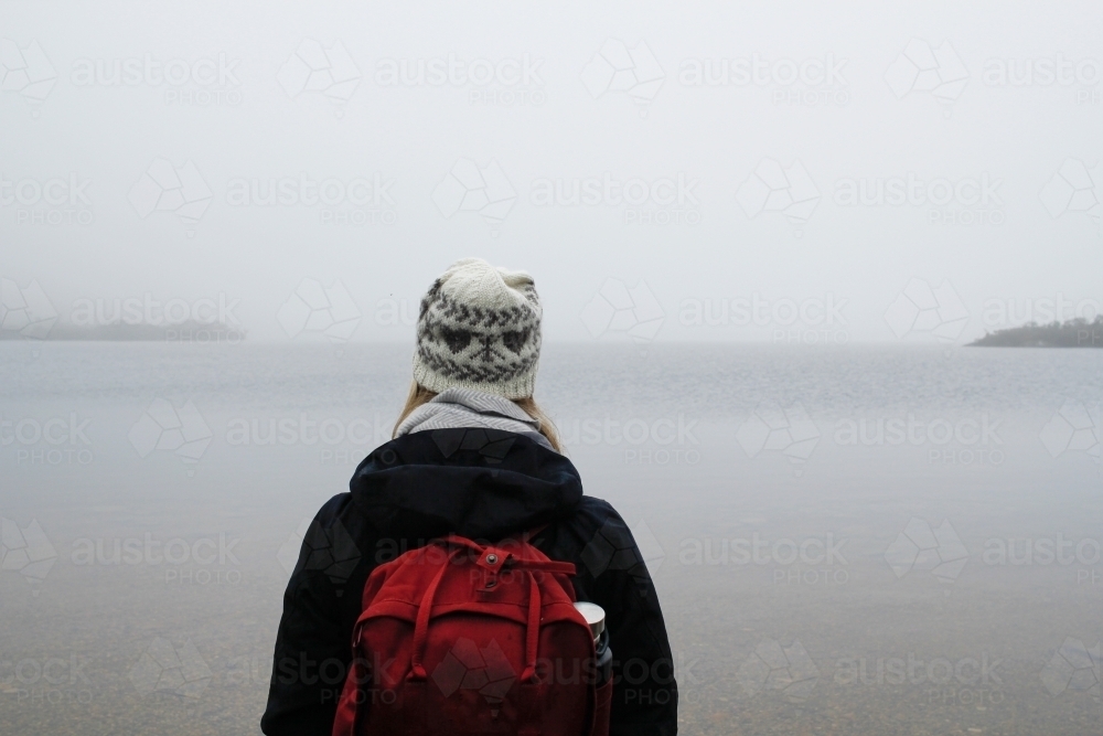 Woman wearing beanie, jacket and backpack looking out over a lake - Australian Stock Image
