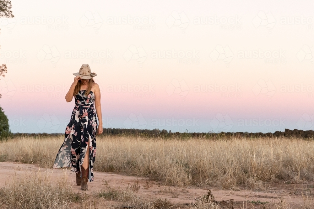Woman wearing floral dress cowboy boots and hat while walking on a dirt road in rural area - Australian Stock Image