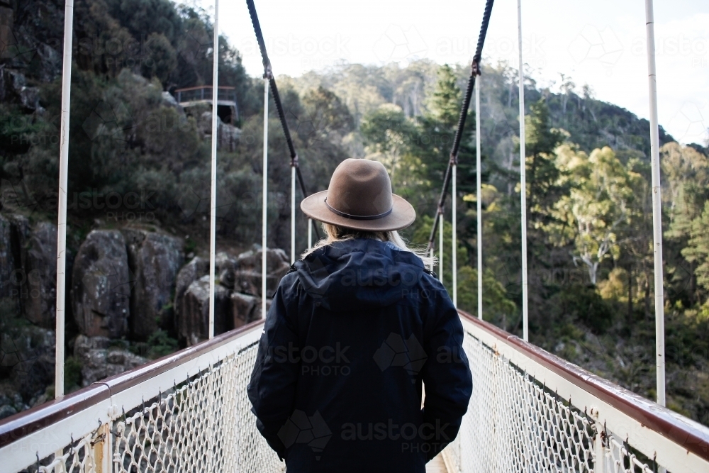 Woman wearing hat and jacket looking out over suspension bridge - Australian Stock Image