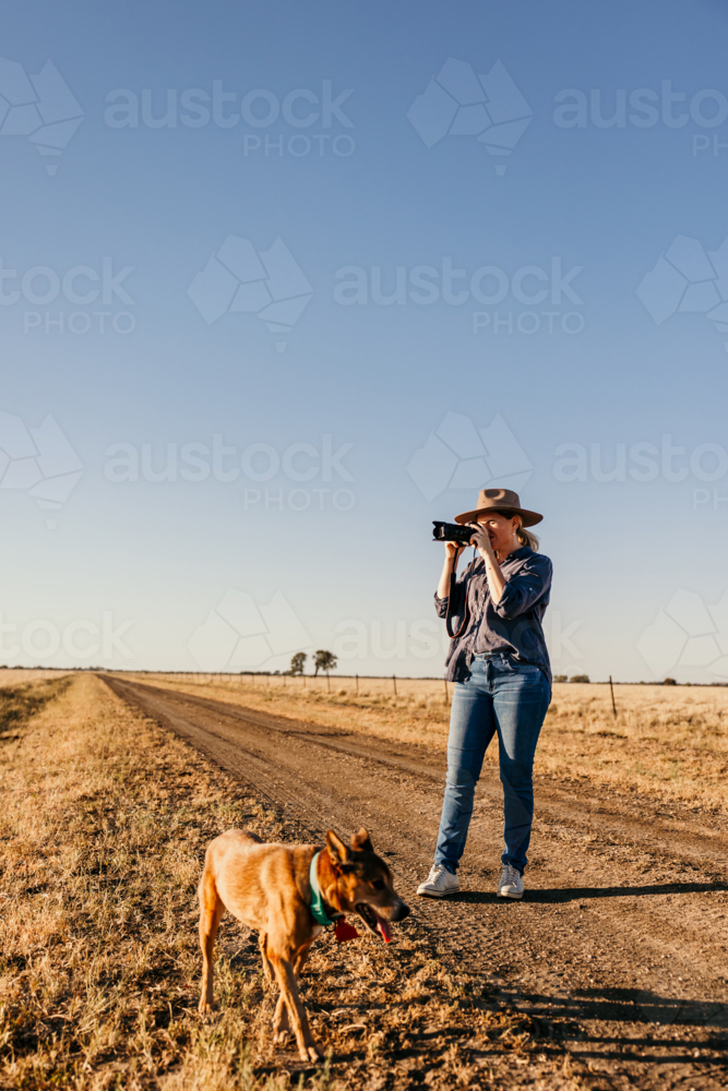 Woman wearing hat standing on the dirt road taking pictures. - Australian Stock Image