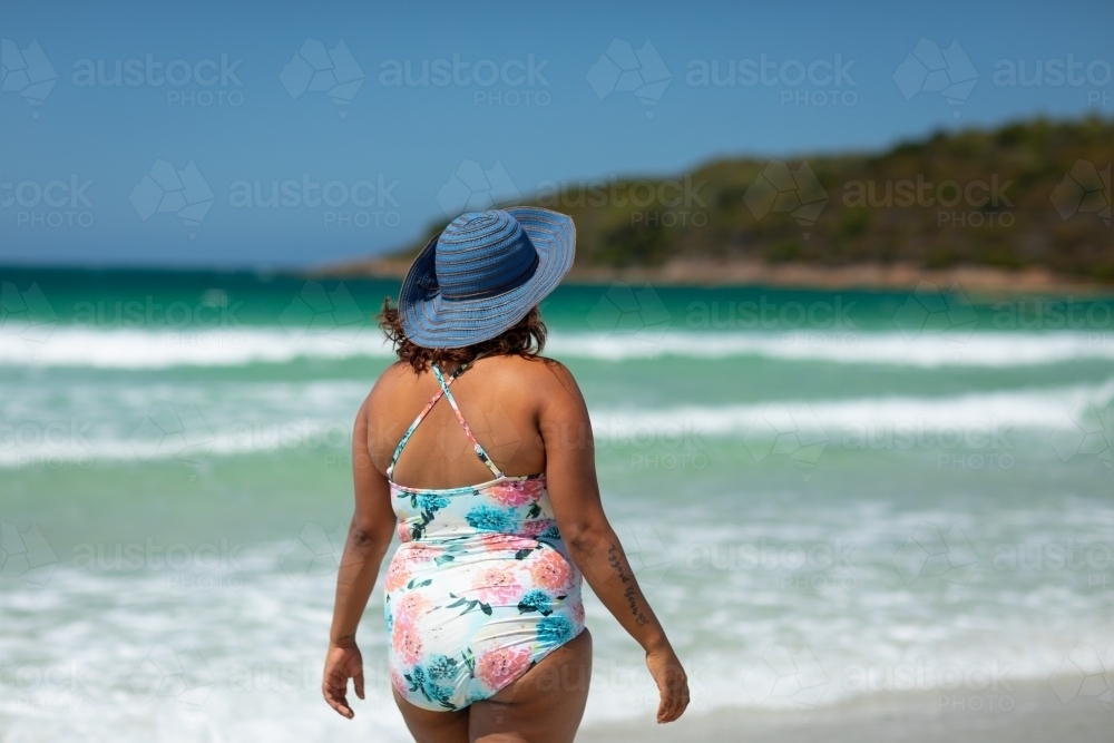 woman wearing sunhat and swimsuit on beach seen from behind - Australian Stock Image